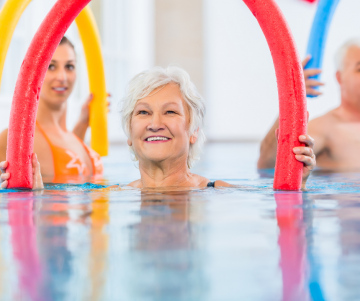 Schwimmschule Swim2grow: Aquafitness für Senioren: Auf dem Bild ist eine Seniorin im Wasser zu sehen. In der Hand hält sie eine rote Nudel, die kreisförmig aus dem Wasser herausragt. Sie lächelt in die Richtung des Betrachters. Hinter ihr ist eine jüngere