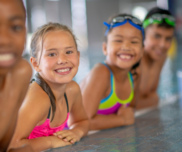 4 Kinder in Schwimmkleidung stützen sich auf den Beckenrand und lächeln in die Kamera. Die zwei hinteren Kinder tragen jeweils eine Schwimmbrille.
