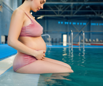 Schwangerenschwimmen bei der swim2grow: Zu sehen ist eine Frau, deren Füße in das Wasser hängen, während sie sitzt. Sie streichelt ihren Bauch und blickt auf diesen.