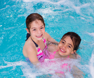 Zwei Mädchen halten sich im Wasser im Arm und lächeln in die Kamera. Beide tragen einen pinken Badeanzug. Um sie herum sind viele Blubberbläschen vom Wasser.