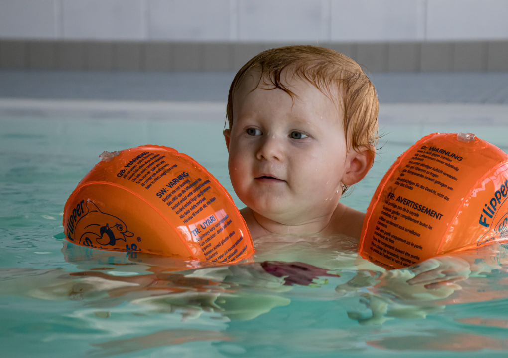 Eine Mutter hält von hinten ihr Baby im Arm, dieses blickt und lächelt in die Richtung des Betrachters. Beide sind im Wasser.