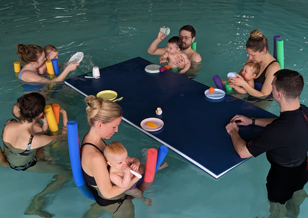 Ein Mutter und ihr Baby sind in einem Schwimmbecken. Die Mutter schaut der Kleinen ins Gesicht, während das Baby lächelt.