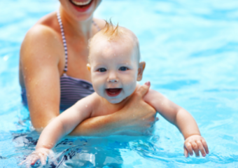 Babyschwimmen Eine Mutter hält von hinten ihr Baby im Arm, dieses blickt und lächelt in die Richtung des Betrachters. Beide sind im Wasser.