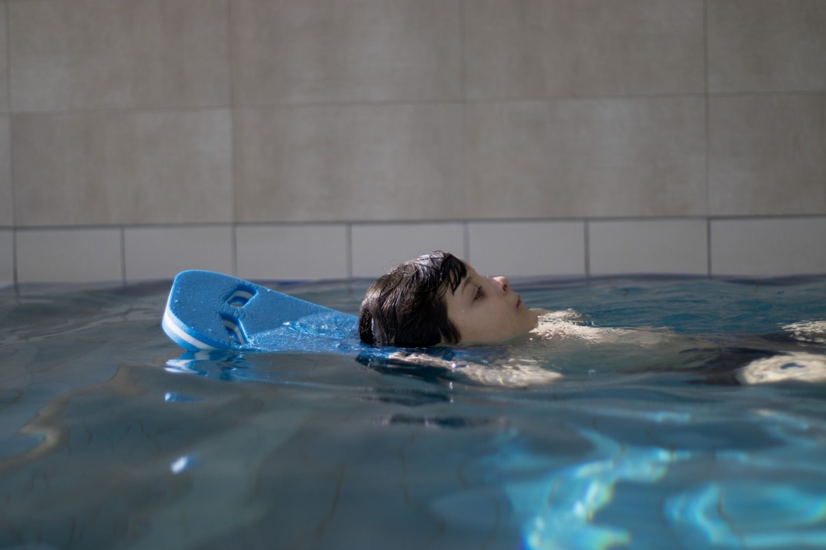 Zwei Mädchen, beide tragen eine Schwimmbrille, stehen im Wasser und sind an den Beckenrand gelehnt. Sie blicken sich in die Augen, während sie sich unterhalten.