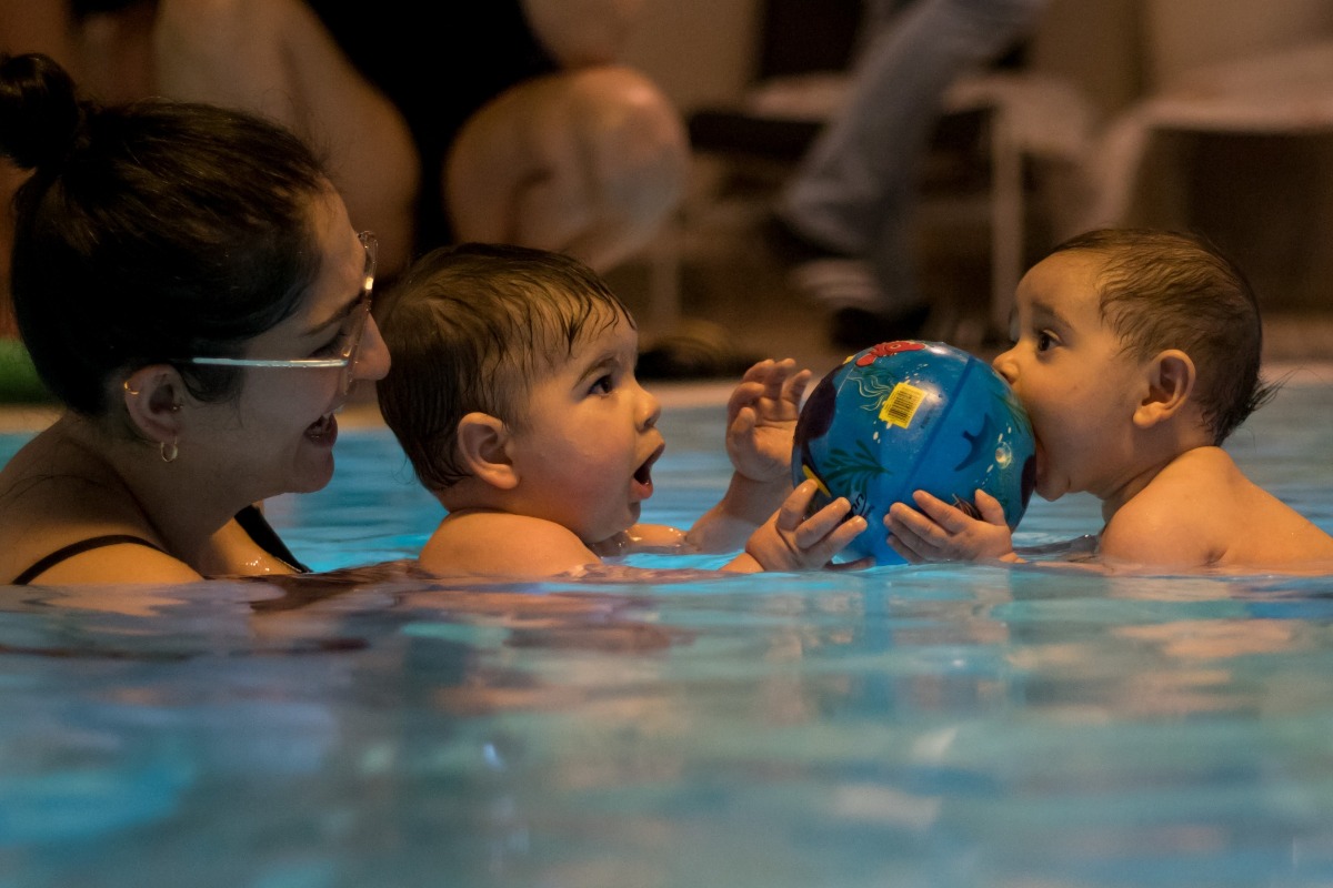 Eine Mutter hält von hinten ihr Baby im Arm, dieses blickt und lächelt in die Richtung des Betrachters. Beide sind im Wasser.