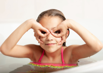 Kinderschwimmen Ein Mädchen mit Badeanzug sitzt in der Wanne und hält ihre Hände vor das Gesicht, sodass es aussieht als würde sie eine Maske tragen.