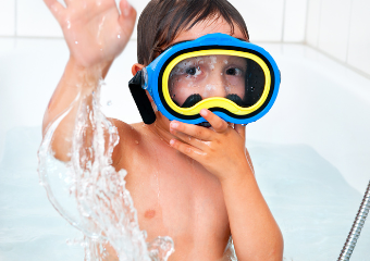 Ein Kind mit braunen Haaren sitzt in der Badewanne und schaut in die Kamera, er trägt eine Tauchermaske mit zwei Farben und spritzt mit der einen Hand Wasser in Richtung Kamera, mit der anderen Hand drückt er die Kamera fest auf sein Gesicht. 