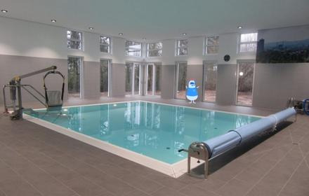 Moderner Raum, in der Mitte das Schwimmbecken der Reha mit Hilfe zum EInstieg, dahinter, hohe Fenster