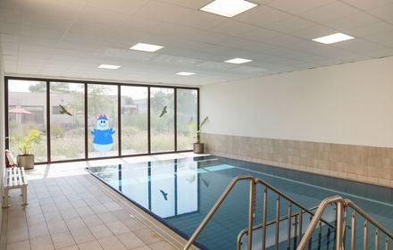 Beckenrand mit weißen Fließen, in der Mitte das Schwimmbecken des Reha-Zentrums in einem hellen Raum, im Hintergrund eine große Fensterwand mit Blick ins Grüne