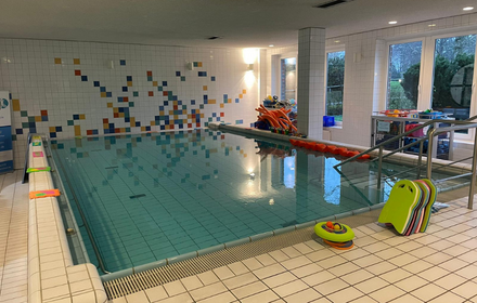 Schwimmbecken, am Beckenrand stehen bunte Schwimmbretter aufgereiht sowie weitere Materialien für den Schwimmunterricht, im Hintergrund eine weiße Wand mit bunten Farbtupfern