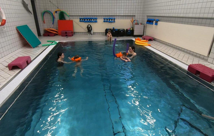 Beim Schwimmunterricht helfen zwei Schwimmlehrerinnen den Kindern beim Beine Schwimmen, die Kinder halten ein Brettchen in der Hand, Schwimmflügel und Nudel tragen sie