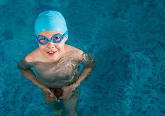 Ein Junge im Wasser trägt eine hellblaue Bademütze sowie eine hellblaue Schwimmbrille.
