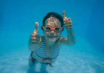Ein Mädchen taucht unter Wasser, trägt eine orangene Schwimmbrille und zeigt beide Daumen nach oben.