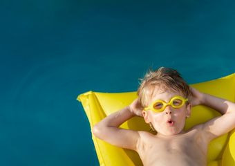 Ein Kind mit gelber Brille auf den Augen, liegt auf einer gelben Luftmatratze im Wasser und formt seine Lippen zu einem "o". Seine Hände sind hinter seinen Kopf gelegt.