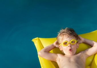 Individualität der Kinder beim Schwimmen Ein Kind mit gelber Brille auf den Augen, liegt auf einer gelben Luftmatratze im Wasser und formt seine Lippen zu einem "o". Seine Hände sind hinter seinen Kopf gelegt.
