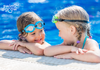 Zwei Mädchen, beide tragen eine Schwimmbrille, stehen im Wasser und sind an den Beckenrand gelehnt. Sie blicken sich in die Augen, während sie sich unterhalten.