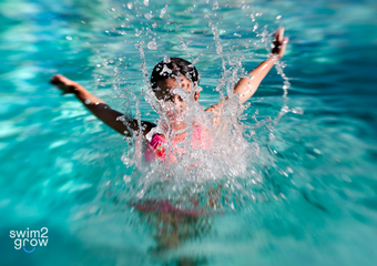 Schwimmabzeichen Seepferdchen Ein Kind im Wasser, das mit den Händen auf das Wasser klatscht, sodass um es herum sehr viele Wasserspritzer sind.