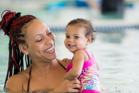 eine Mama lächelt ihr Kind an, welches gerade viel Spaß im Wasser hat