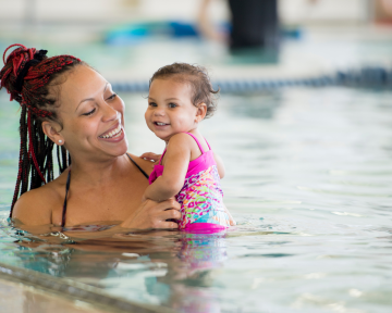Beim Babyschwimmen haben auch unsere Kinder von 12-23 Monaten viel Freude im Wasser eine Mama lächelt ihr Kind an, welches gerade viel Spaß im Wasser hat