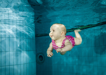 Ein Baby in pinkem Badeanzug taucht unter Wasser und hält seine Augen geöffnet.