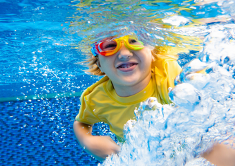 Ein Junge in einem gelben Schwimmshirt mit bunter Brille unter Wasser, vor ihm wirbelt sich das Wasser auf.