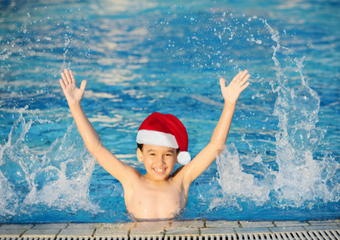 Ein Junge im Wasser, mit der Brust an den Beckenrand gelehnt, streckt seine Arme in die Luft und trägt im Wasser eine Nikolausmütze.