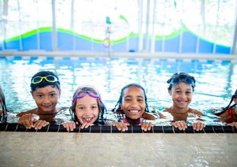Mehrere Kinder im Schwimmbecken, halten sich am Beckenrand und lächeln. 