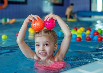 Ein Mädchen im Schwimmbecken, hält sich zwei Spielbälle über dem Kopf, im Hintergrund sehr viele bunte Bälle im Wasser.